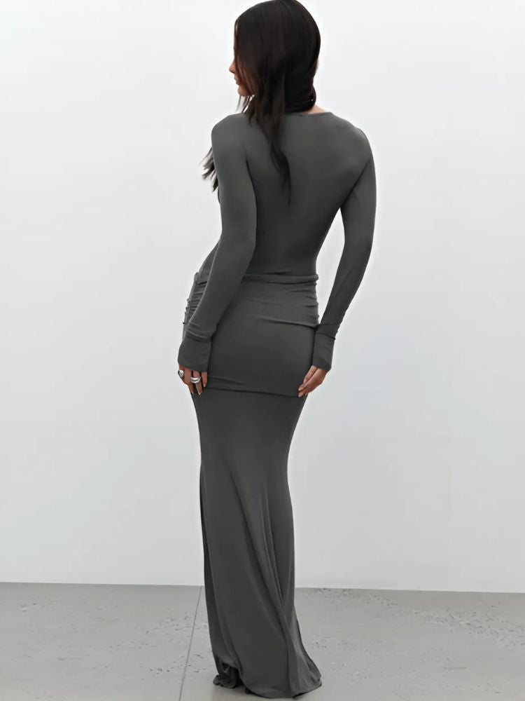 A woman wearing a grey long-sleeved bodycon maxi dress with diagonal ruched details on the chest and hip, featuring a fitted silhouette and a flowy hem from the knee down.