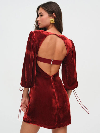 Woman wearng a deep red velvet mini dress. The dress features a structured bust, long billowy sleeves with adjustable ties, and an open back with a clasp.