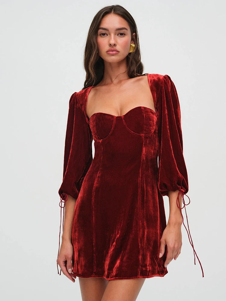 Woman wearng a deep red velvet mini dress. The dress features a structured bust, long billowy sleeves with adjustable ties, and an open back with a clasp.