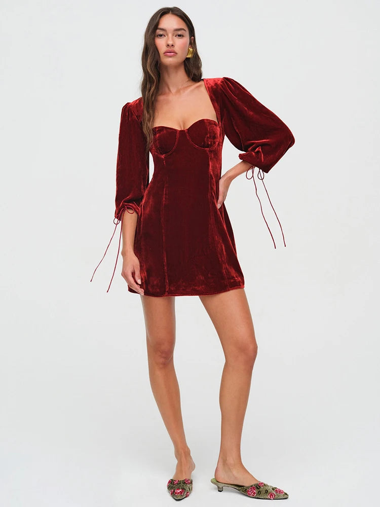 Woman wearng a deep red velvet mini dress. The dress features a structured bust, long billowy sleeves with adjustable ties, and an open back with a clasp.