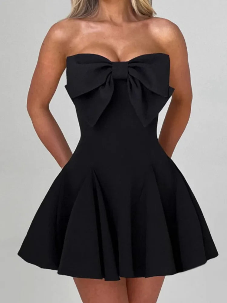 Woman wearing a black strapless and backless Mini Dress featuring an oversized bow on the front, an A-line silhouette, and pleated skirt