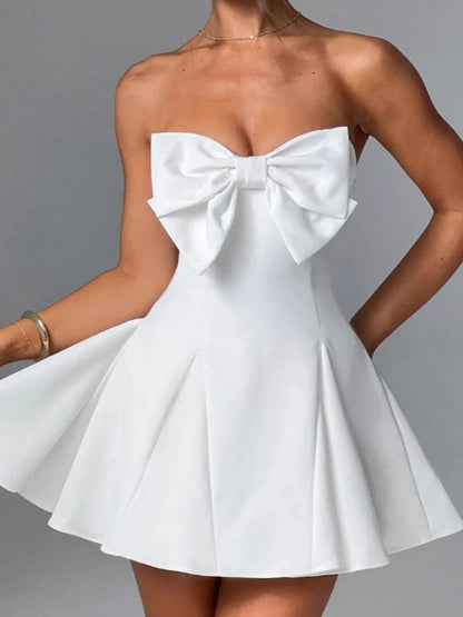 Woman wearing a white strapless and backless Mini Dress featuring an oversized bow on the front, an A-line silhouette, and pleated skirt