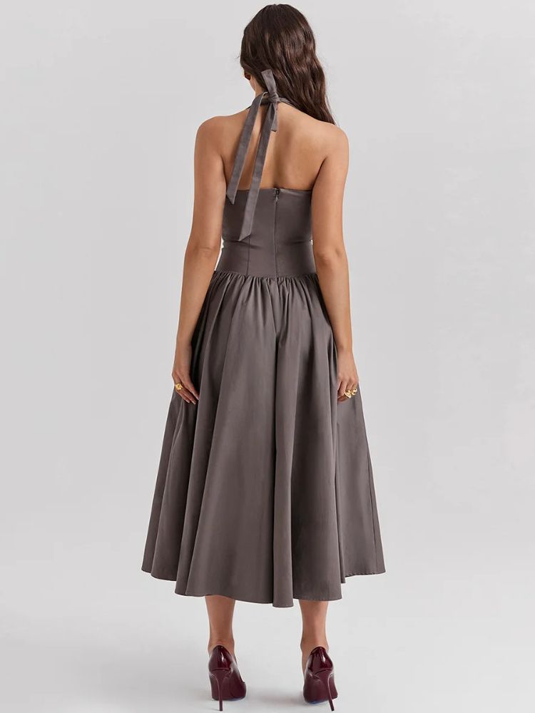 Woman wearing a khaki gray midaxi dress with a deep halter v neck, pleated skirt an neck mounted backless design.