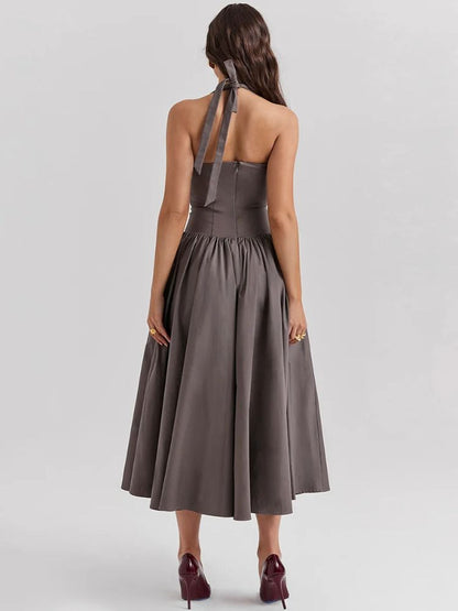 Woman wearing a khaki gray midaxi dress with a deep halter v neck, pleated skirt an neck mounted backless design.