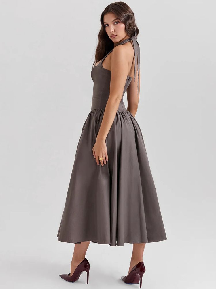 Woman wearing a khaki gray midaxi dress with a deep halter v neck, pleated skirt an neck mounted backless design.