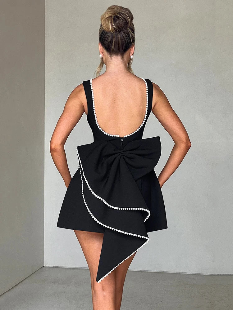 Woman wearing a black backless mini dress with pearl bow detail and pearl-embellished collar, sleeveless high-waist A-line silhouette.